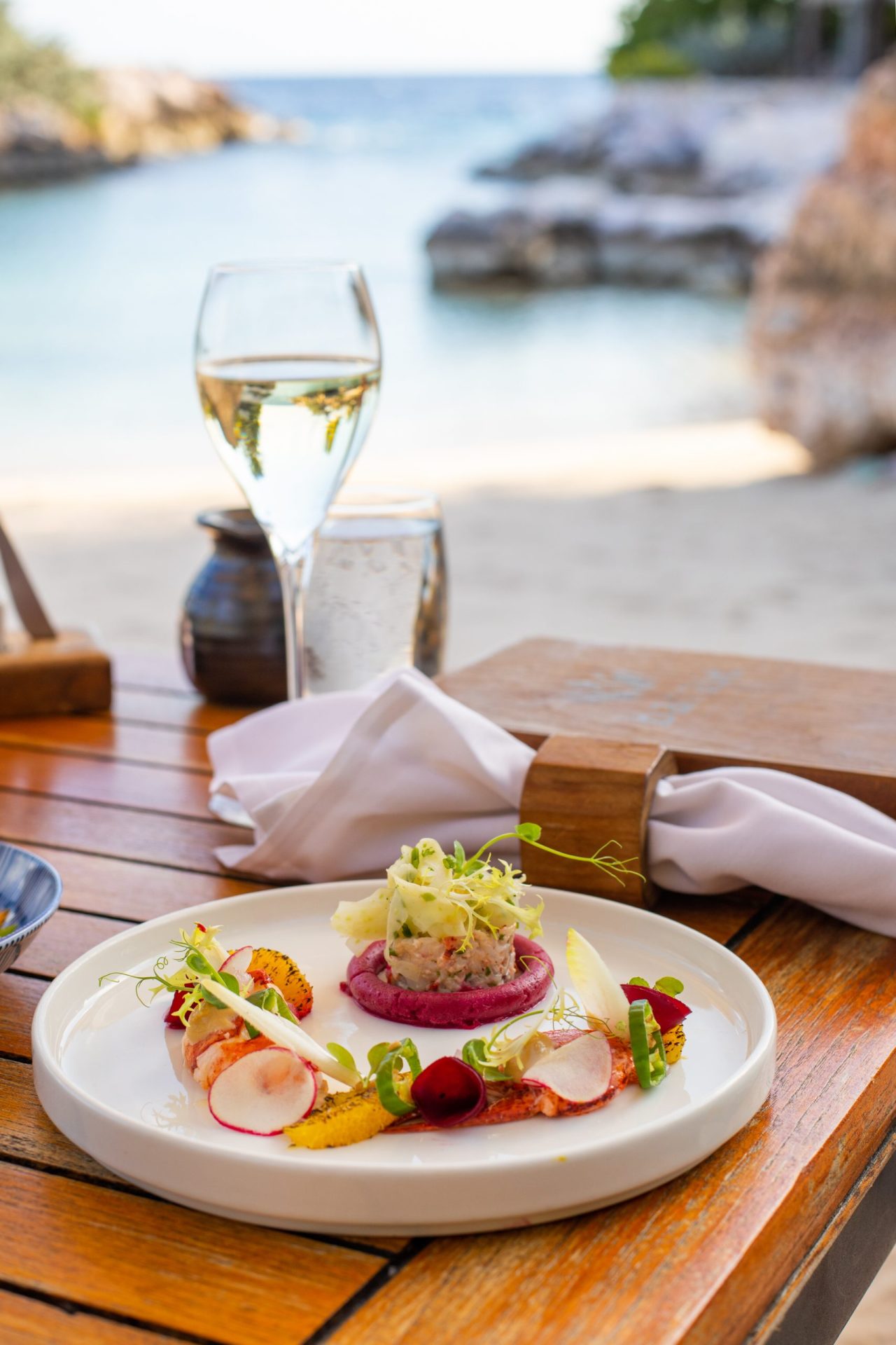 A plate of food and a glass of wine on a table with the beach in the background